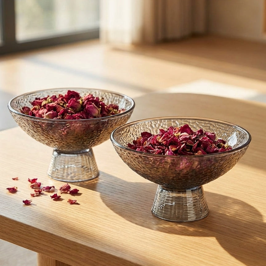 Two glass bowls with dried flowers on a wooden table near a window.