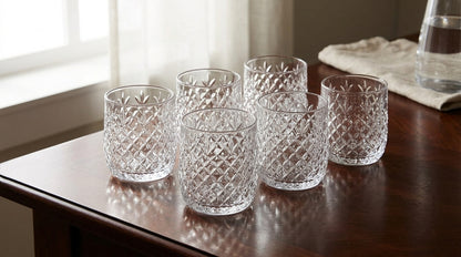 Five clear cut-crystal tumbler glasses arranged on a dark wooden table in a softly lit room, with a window and curtain in the background.