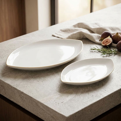 Two white ceramic serving platters with a thin gold rim, displayed on a light gray stone countertop with figs, rosemary, and a linen napkin.
