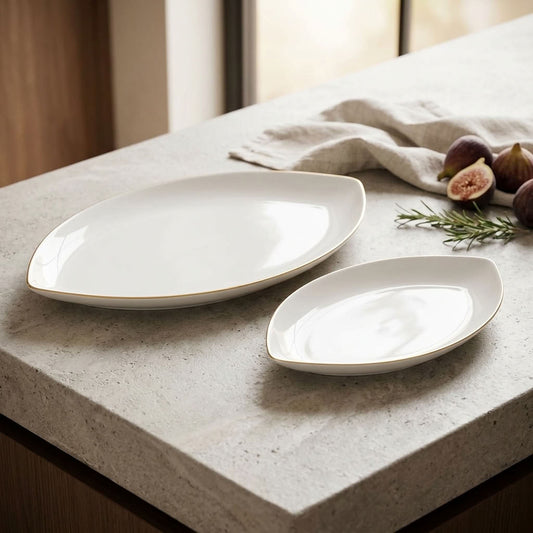 Two white ceramic serving platters with a thin gold rim, displayed on a light gray stone countertop with figs, rosemary, and a linen napkin.