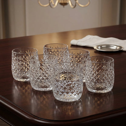 Five clear cut-crystal tumblers arranged on a dark wooden table in a dining room setting, with a chandelier and folded white napkin visible in the background.