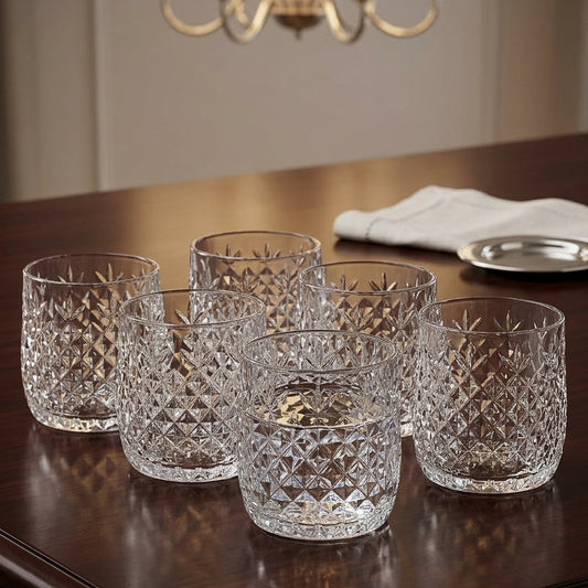 Five clear cut-crystal tumblers arranged on a dark wooden table in a dining room setting, with a chandelier and folded white napkin visible in the background.