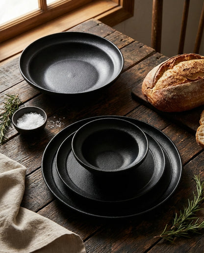 Set of black ceramic plates on a wooden table with bread and herbs.