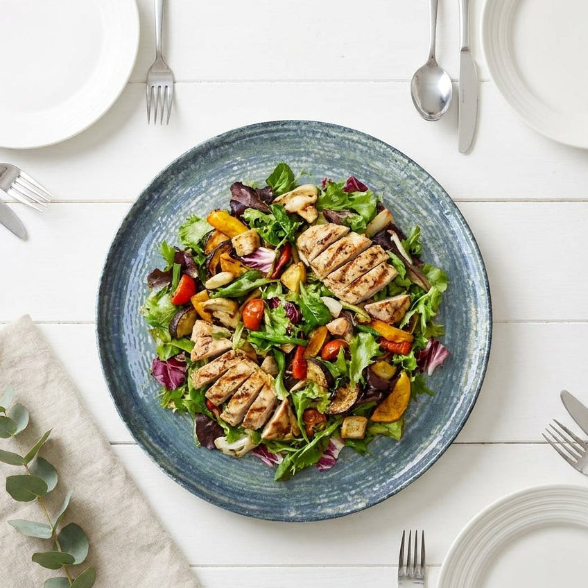 Salad with grilled chicken and vegetables on a blue plate, set on a white table.