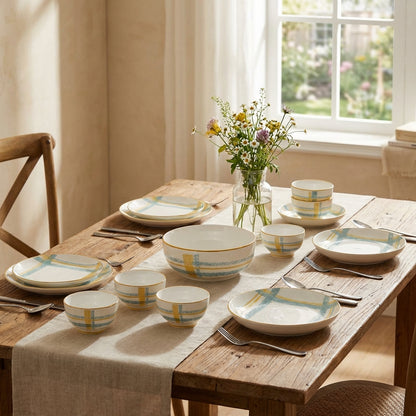 Dining table set with ceramic plates, bowls, and cutlery on a wooden table with a window in the background.