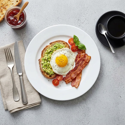 A styled breakfast scene on a light gray surface showing a white plate with avocado toast, a sunny-side-up egg, bacon, cherry tomatoes, and basil. A fork, knife, beige napkin, jam jar, and black coffee cup on a saucer are arranged around it.