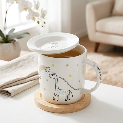 A white ceramic mug with a matching lid, featuring a cute giraffe illustration, a small rainbow, and colorful polka dots, sitting on a wooden coaster on a white table in a bright, modern room.