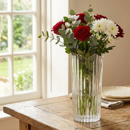 A clear ribbed glass vase filled with a bouquet of red roses, white daisies, and green eucalyptus leaves, set on a rustic wooden table in front of a softly lit window.