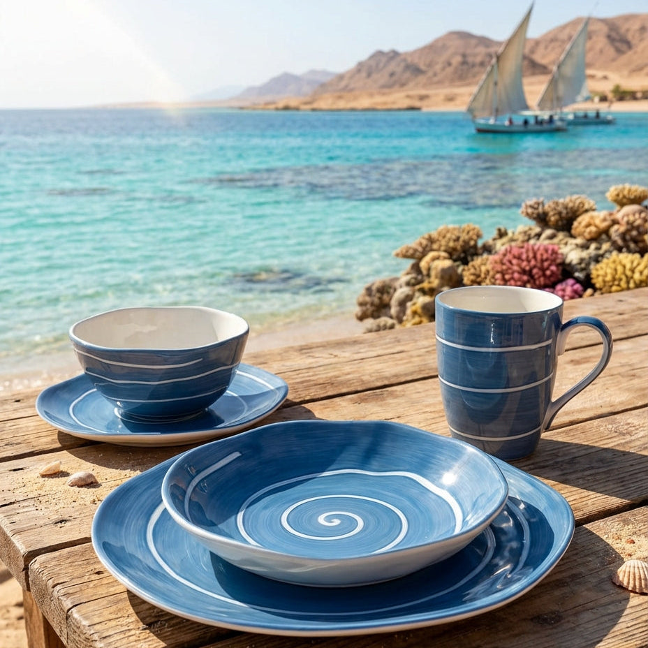 Blue ceramic tableware set on a wooden table with a scenic ocean view and sailboat.