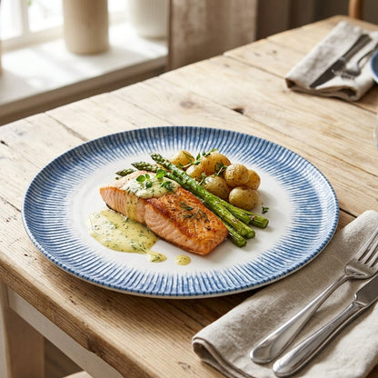 A round white plate with a blue ribbed rim, styled on a rustic wooden table with a salmon fillet, asparagus, potatoes, and a green sauce, alongside a folded beige napkin and silver fork and knife.