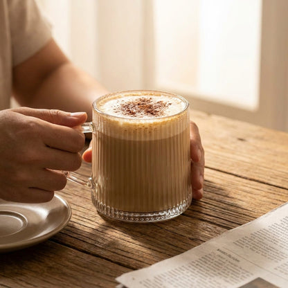 Person holding a glass mug of coffee with a wooden table and newspaper in the background