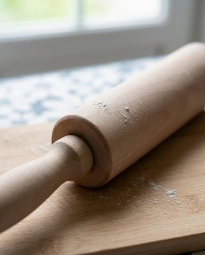 A light natural wood rolling pin resting on a matching wooden cutting board with a few scattered flour particles, set against a softly blurred kitchen background near a window.