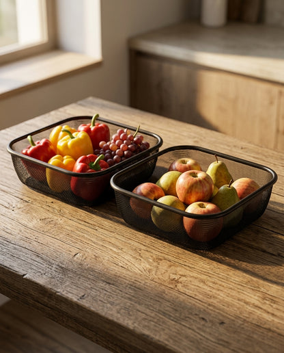 Two black mesh rectangular baskets filled with fruit, including apples, pears, grapes, and bell peppers, arranged on a rustic wooden table in a sunlit kitchen setting.