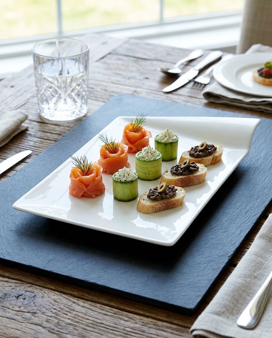 A white rectangular serving platter holding six gourmet appetizers, including smoked salmon, cucumber wheels, and small bread slices with dark spread, set on a dark slate board on a rustic wooden table.