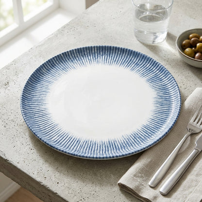 A round ceramic plate with a white center and a blue ribbed rim, styled on a speckled stone table with a fork, napkin, glass of water, and olives nearby.