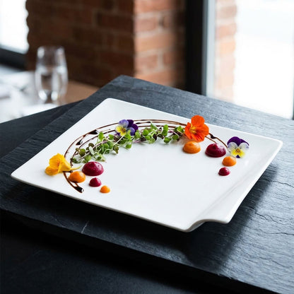 A white rectangular ceramic serving platter with rounded corners, styled with edible flowers, greens, and sauce on a dark slate table in a restaurant setting.