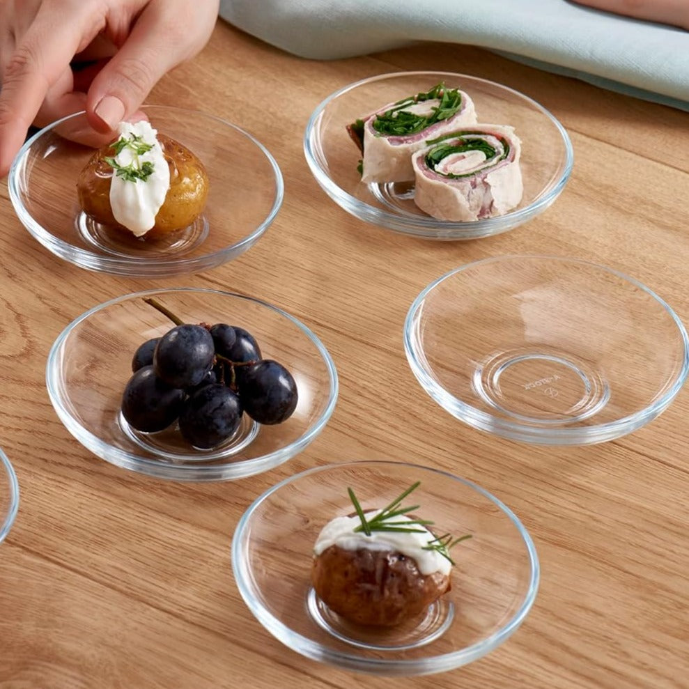 Clear glass saucers with various dishes served on them, including a rolled-up pastry, a slice of cake, a ball of ice cream, a slice of pizza, a cluster of grapes, and a dish with a green herb topping.