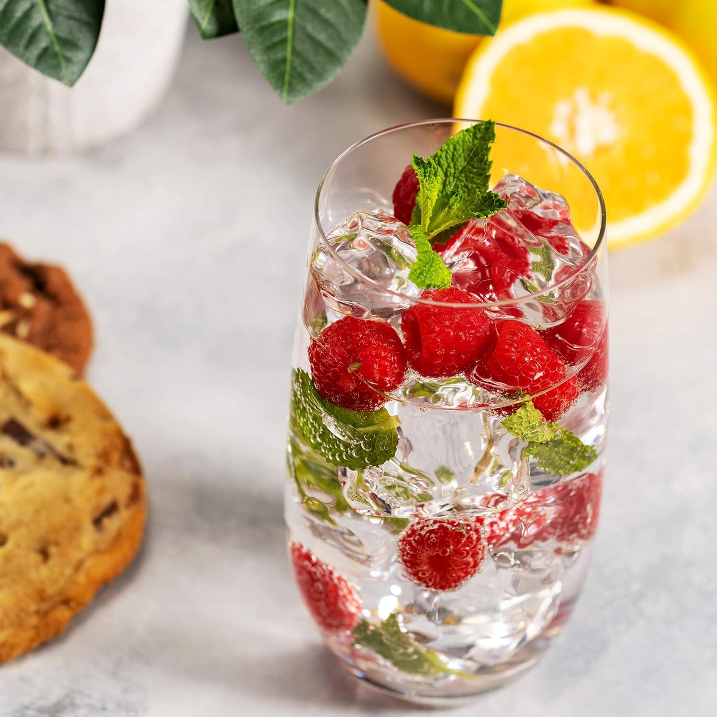 A clear highball glass filled with water and raspberries, accompanied by a cookie and a slice of lemon on a grey surface.