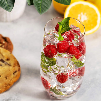 A clear highball glass filled with water and raspberries, accompanied by a cookie and a slice of lemon on a grey surface.