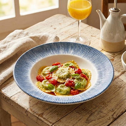 A round ceramic plate with a blue ribbed rim and white interior, filled with ravioli, green sauce, and red tomatoes, set on a rustic wooden table with a glass of orange juice and a cream-colored teapot in the background.