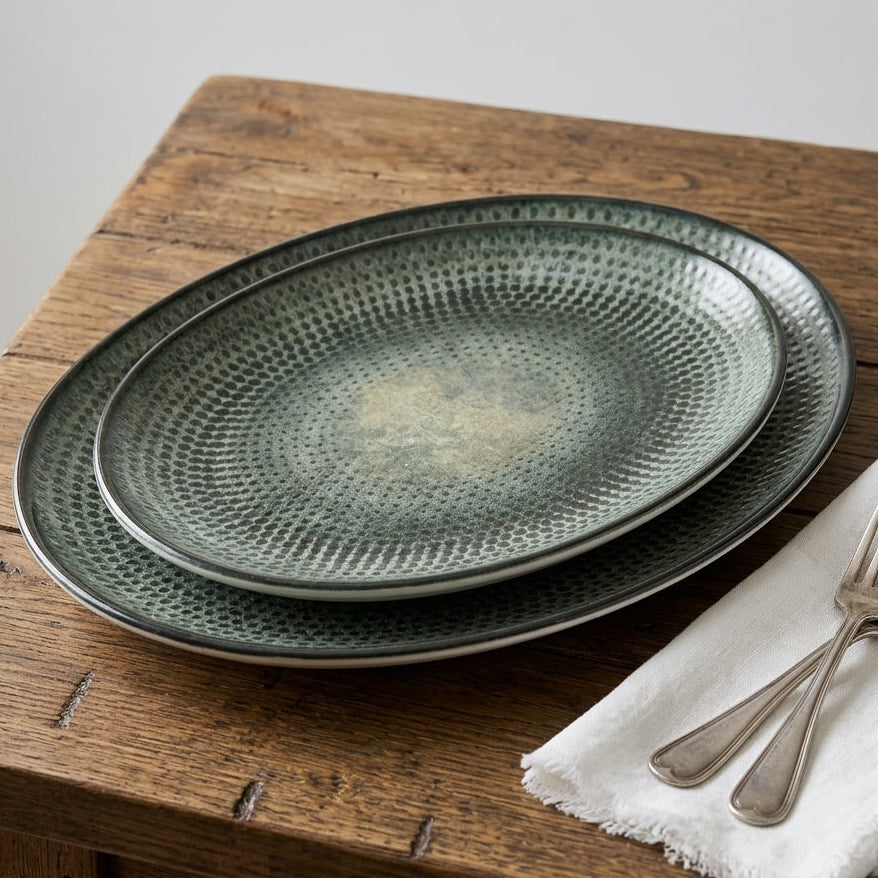 Two oval ceramic plates with a textured, hammered green glaze, stacked on a rustic wooden table next to a white napkin and silver forks.