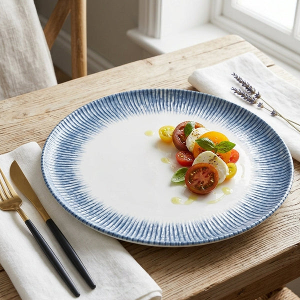 A round white ceramic plate with a blue hand-painted radial pattern around the rim, styled on a rustic wooden table with a salad of tomatoes and mozzarella, alongside gold and black cutlery on white napkins.