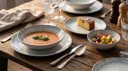A rustic dining table set with a bowl of tomato soup garnished with basil, a plate of fruit salad, a slice of cake, and matching speckled gray ceramic plates and bowls.