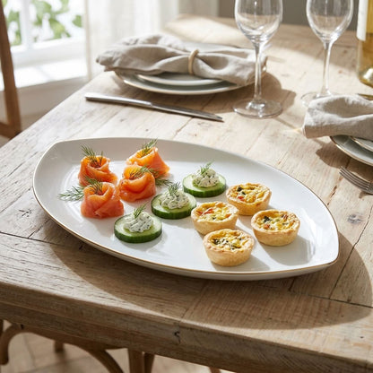 An oval white serving platter with a gold rim, styled on a rustic wooden table with appetizers including salmon, cucumber slices, and tartlets, with a set table in the background.