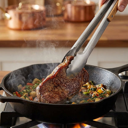 Steak being cooked in a pan with tongs, surrounded by copper pots in a kitchen setting.