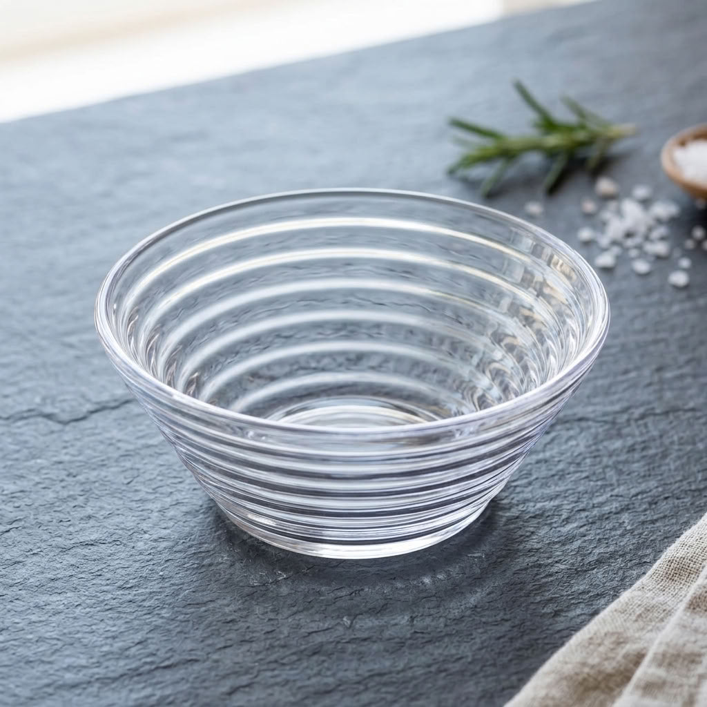 A clear ribbed glass bowl with concentric circular ridges, shown on a dark slate surface with a sprig of rosemary and coarse salt in the background.