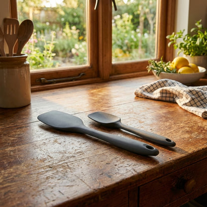 Two black spatulas on a wooden kitchen counter with a window and plants in the background.