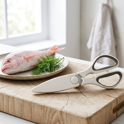 White and gray kitchen shears with a circular pivot, resting on a wooden cutting board next to a plate with a raw fish and dill.
