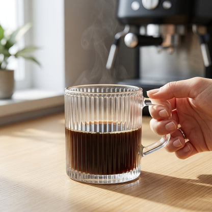 A clear ribbed glass mug filled with dark coffee, held by a hand on a wooden table with a blurred coffee machine and window in the background.