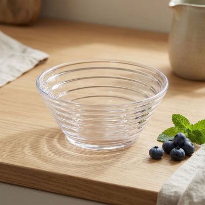 A clear ribbed glass bowl sits on a light wood countertop, with blueberries and mint leaves beside it and a softly blurred kitchen background.