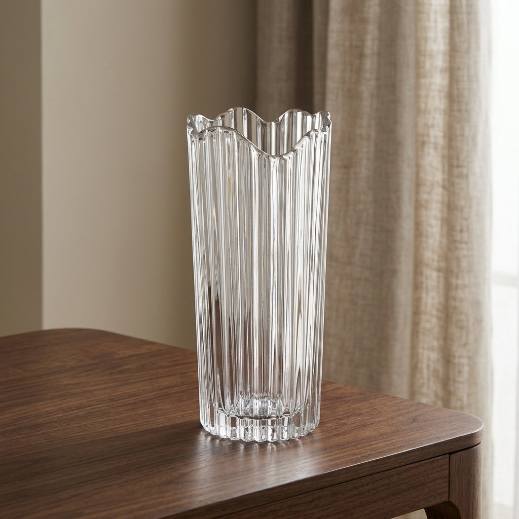 A tall, clear ribbed glass vase with a scalloped rim, standing on a dark wood table in front of beige curtains.