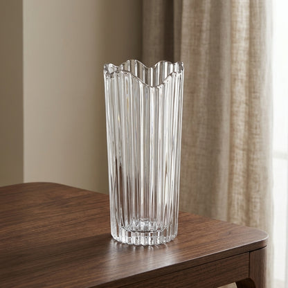 A tall, clear ribbed glass vase with a scalloped rim, standing on a dark wood table in front of beige curtains.