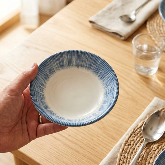 A hand holds a blue and white ceramic bowl with a textured blue rim, set against a warm wooden dining table with place settings and silverware.