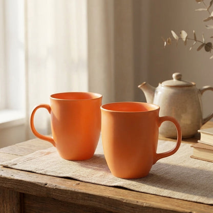 Two orange mugs on a wooden table with a teapot and books in the background.