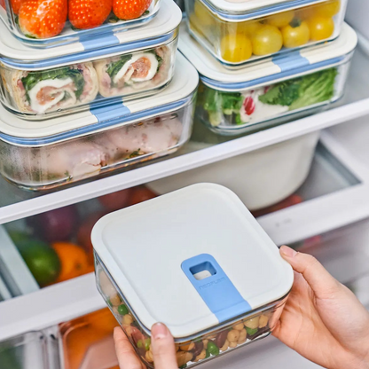 A person holding a square-shaped food storage container with a blue silicone lid, in a refrigerator filled with various other containers.