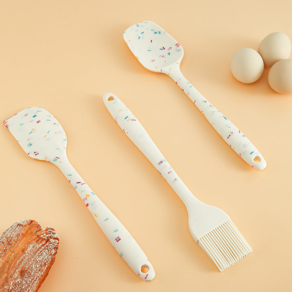 A set of three white silicone kitchen utensils with multicolored speckles, including a spatula, a spoon, and an oil brush, displayed on a beige background with eggs and bread in the peripheral.